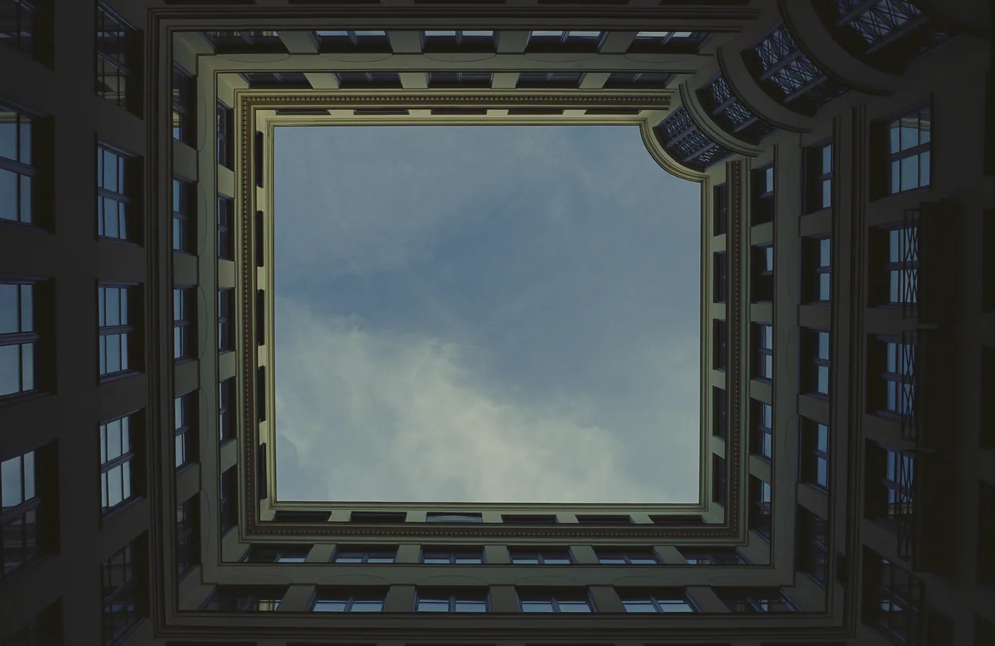 Blick senkrecht nach oben in einen Innenhof, dunkle Fassaden mit Fenstern rahmen ein Stück bewölkten Himmel