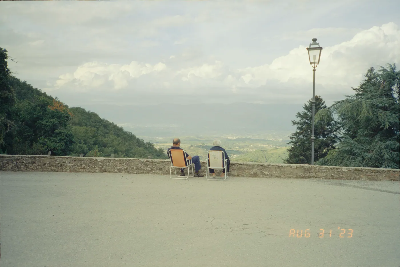 Zwei Personen sitzen in Klappstühlen an einer Steinmauer und blicken über ein weites Tal unter Wolken, rechts eine Straßenlaterne